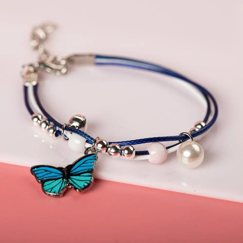 Bracelet avec pendentif papillon bleu perles blanches posé sur fond blanc et rose