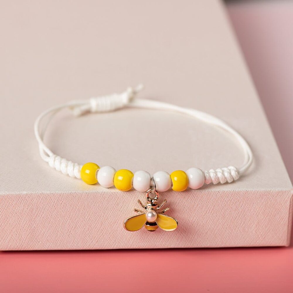 Bracelet tressé blanc avec pendentif abeille et tournesol sur une étagére blanche avec fond rose