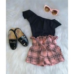 Short rose à carreaux rose avec chemise noire, des lunettes de soleil en rose et des chaussures en noir brillant sur un tapis blanc