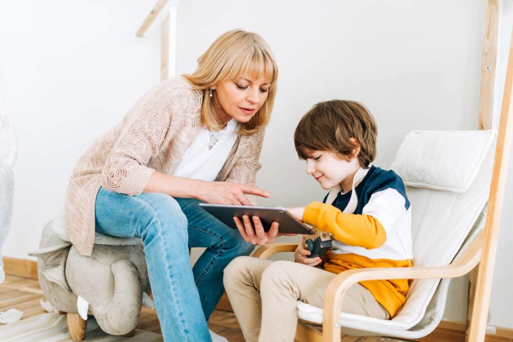 Une mamie qui apprend à lire l'heure à son petit fils sur une tablette ipad. la mamy est blonde avec un gillet gris et un jeans bleu le petit fils a un gilet jaune et bleu et porte un jouet en bois