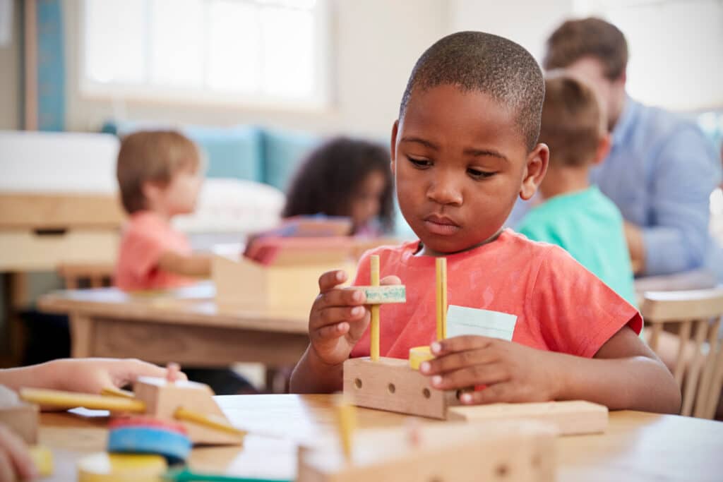 Un petit enfant noir assit à sa table dans sa classe d'école autour d'autres enfants, il joue avec un jouet montessori en bois
