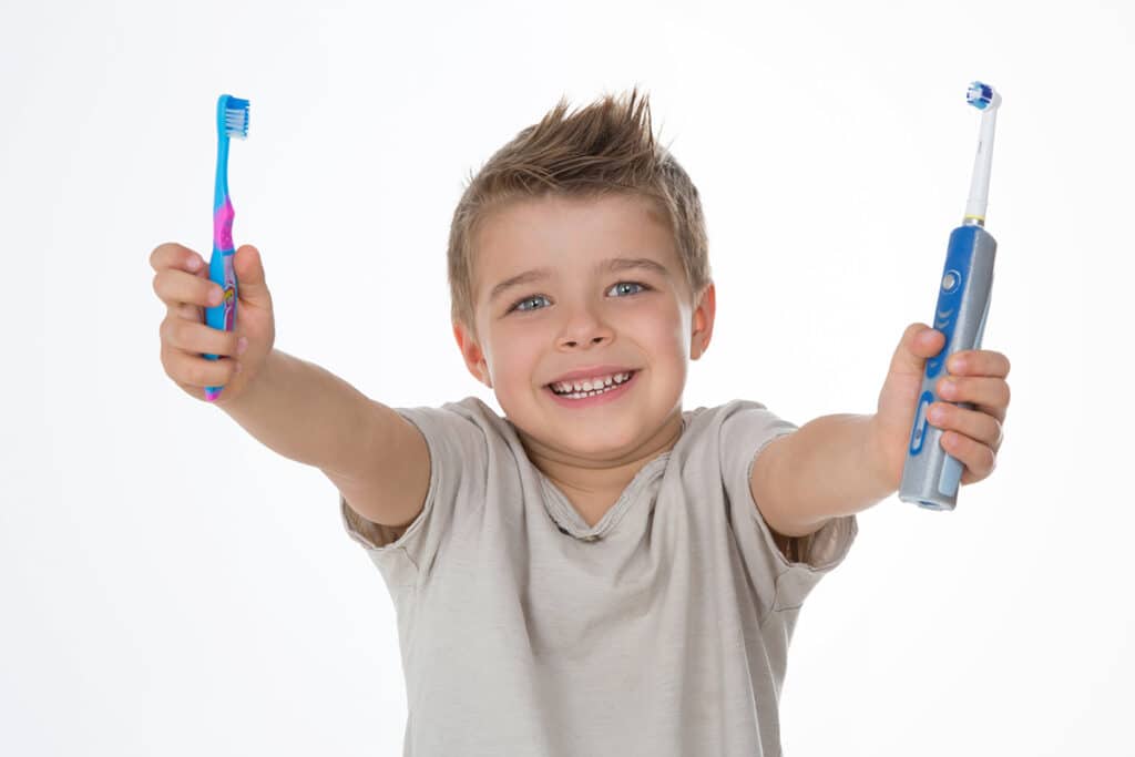 Un petit garçon sur fond blanc qui tient une brosse à dent electrique dans la main gauche et une brosse à dent manuelle dans la main droite. Il porte un t shirt gris