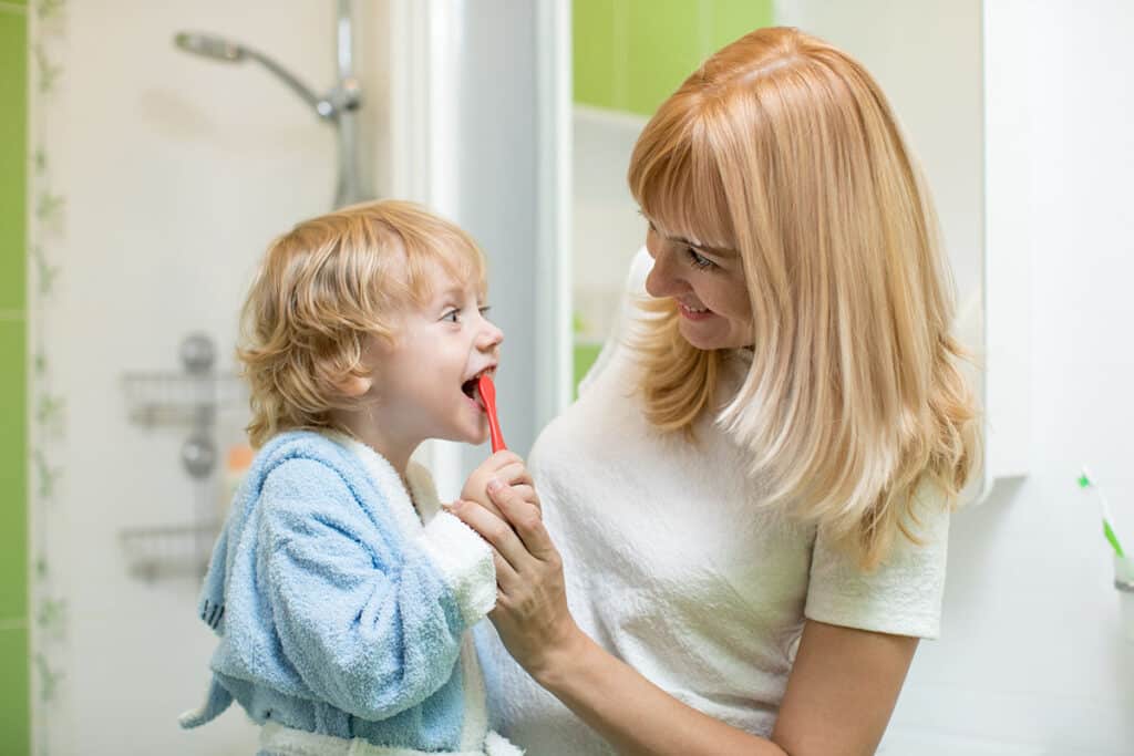 Une maman rousse apprend à son enfant roux à se brosser les dents. la maman porte un t shirt blanc, le petit garçon un peignoir de bain bleu. Ils sont dans la salle de bain.