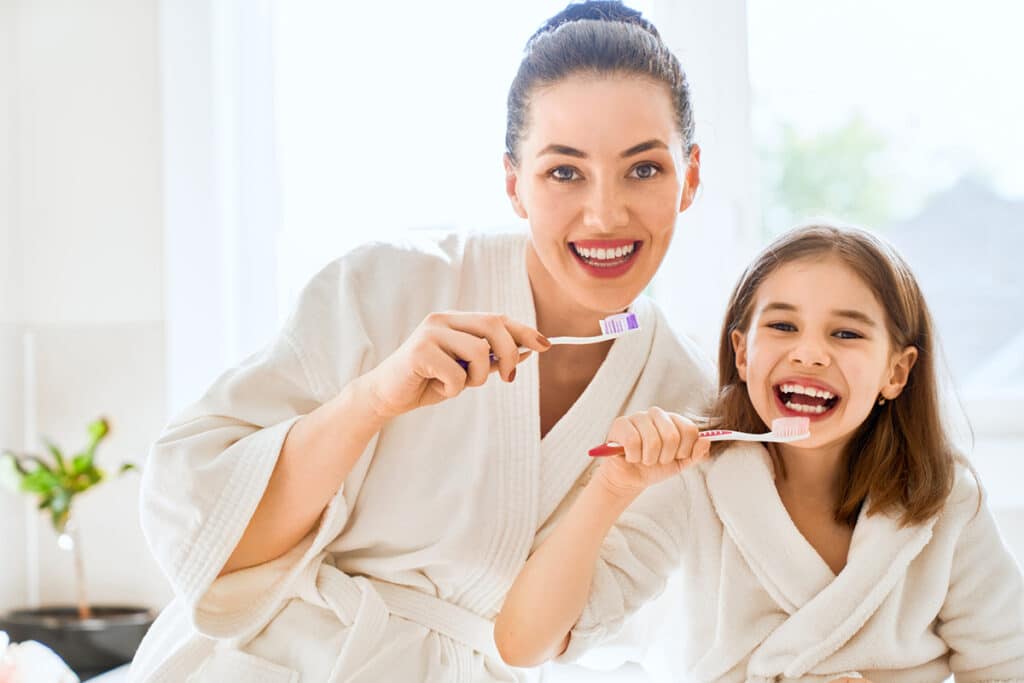 Une maman et sa jeune fille se lavent les dents en souriant. elles portent un peignoir de bain blanc et chacune a une brosse à dent différente , une bleue et une rouge. Il y a une plante verte dans le fond