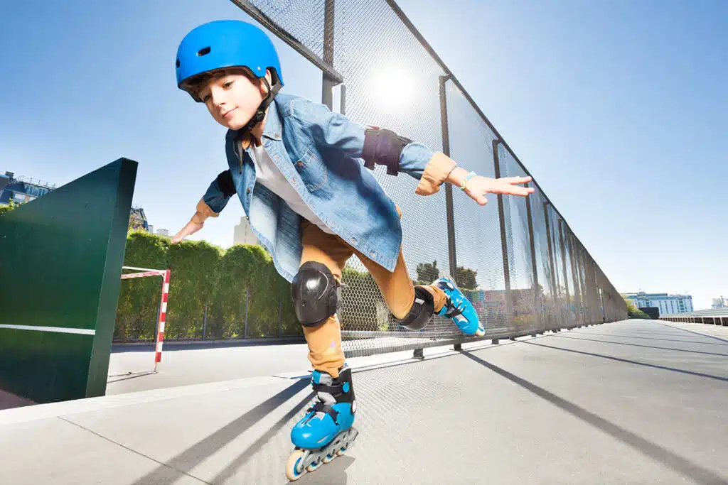 Un jeune garçon qui fait du roller sur le trottoire sous le soleil. Il porte une paires de patins à roulettes bleu, des genouillère noires et des protections aux coudes noires ainsi qu'un casque bleu.