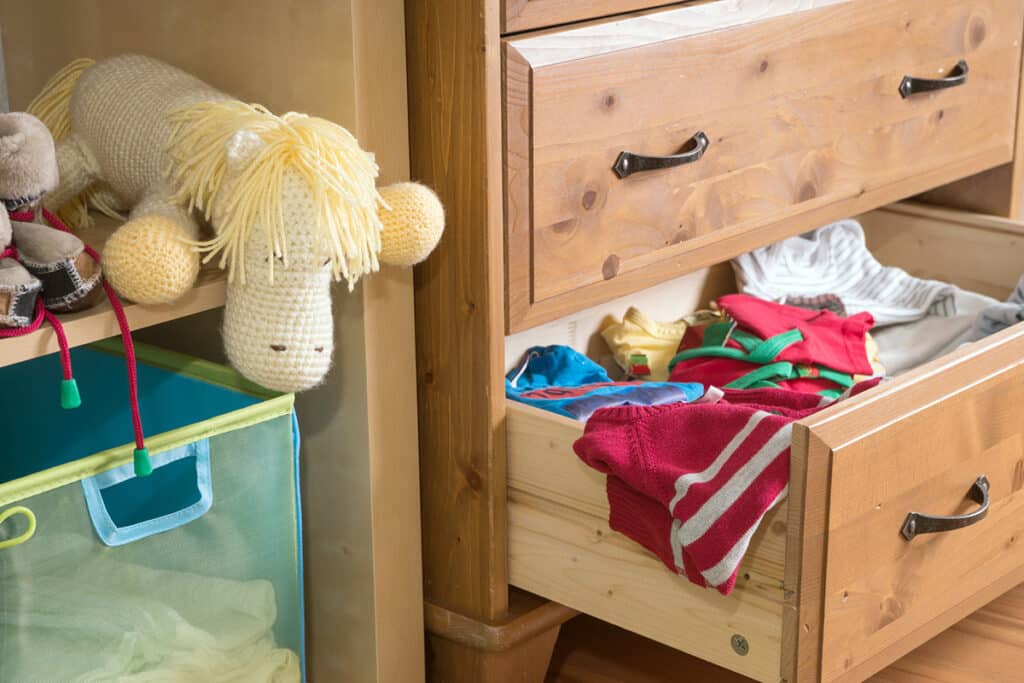 Une armoire en bois avec le dernier tiroir ouvert et des vêtements en désordre en ressortent. à coté il y a une peluche pour enfant blanche et jaune.