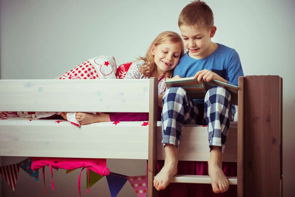 Deux enfants en pyjama sourient en lisant un livre assis sur le lit du haut de leur lit superposé gris. 