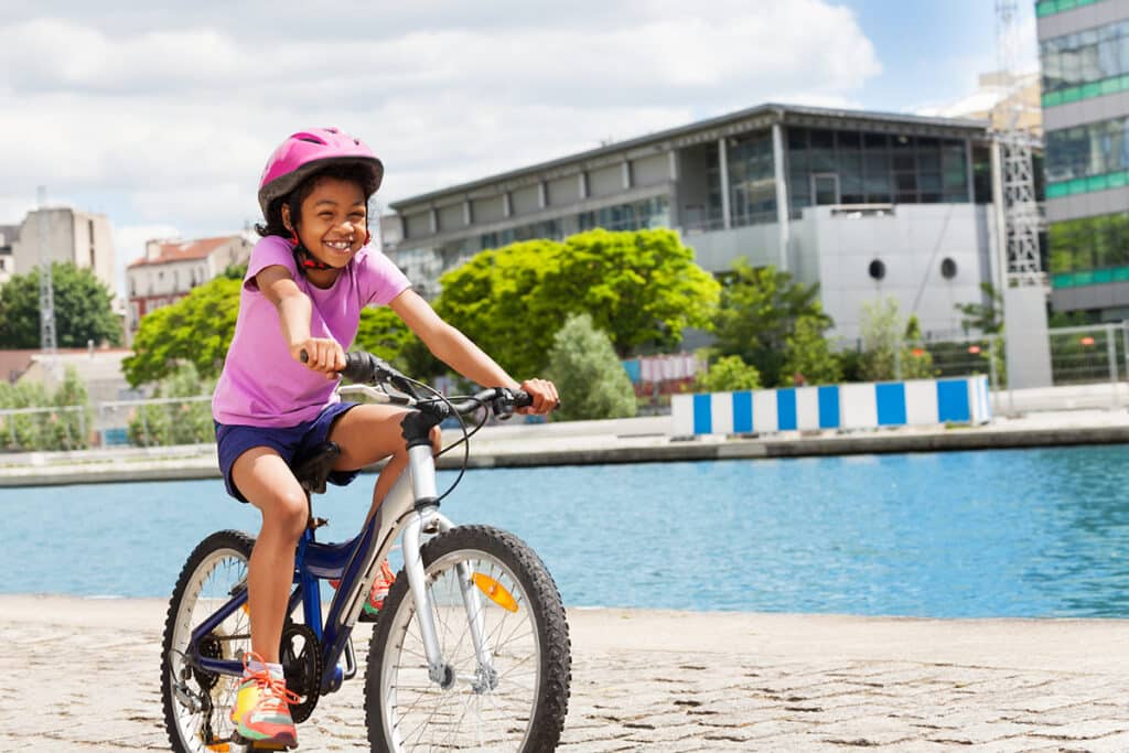 Une jeune fille d'origine afro qui fait du vélo le long du canal en plein air. Elle est vétue d'un t shirt rose, d'un short bleu, d'un casque rose et de sneakers orange. dans le fond on voit des batiments et des arbres verts
