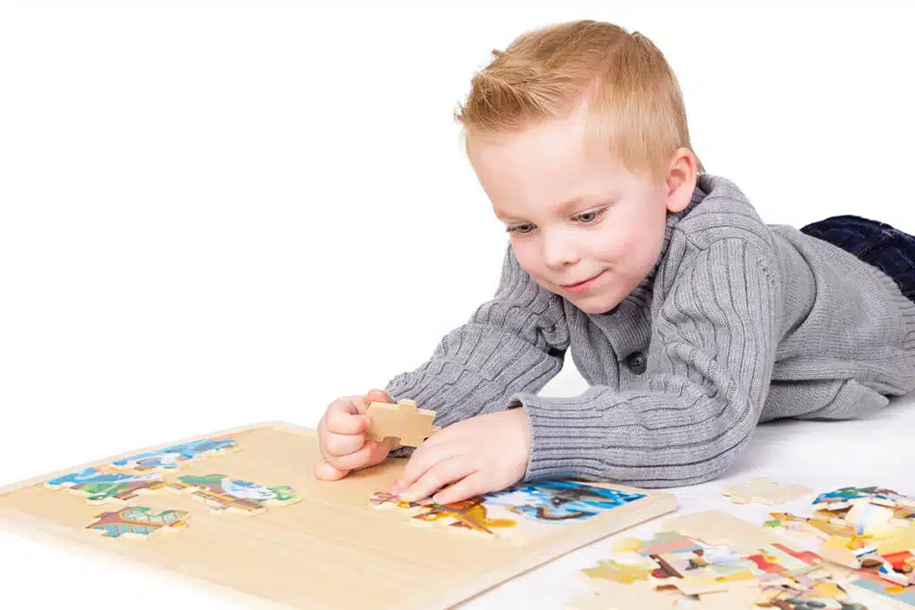 Un petit enfant de 5 ou 6 ans avec des cheveux roux et des yeux bleus. Il est couché par terre et joue avec un puzzle coloré sur fond blanc 
