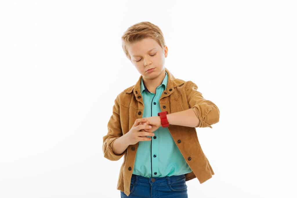 Un jeune garçon sur fond blanc qui lit l'heure sur sa montre connectée, il porte une veste marron et une chemise bleue. 