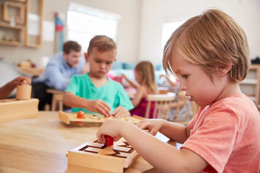 Une jeune fille blonde qui porte un t shirt rose joue avec un jeu montessori en bois dans une classe d'école. En arrière plan il y a d'autres élèves et le professeur d'école