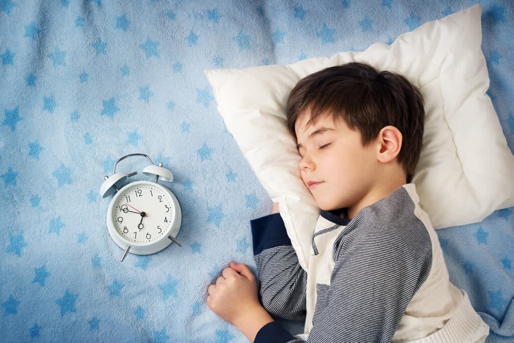 Un jeune garçon endormis dans son lit sur un drap bleu avec des étoiles bleues foncées. Il y a un reveil posé a coté de lui.