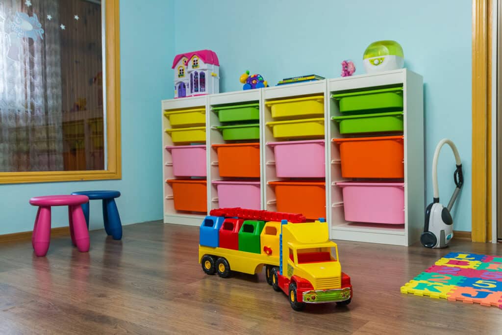 Des meubles de rangement pratiques dans une salle de jeu pour enfant. les meubles ont des bacs de rangement de couleurs. Il y a un jouet camion pour enfant, deux tabourets de couleurs et un tapis de jeu
