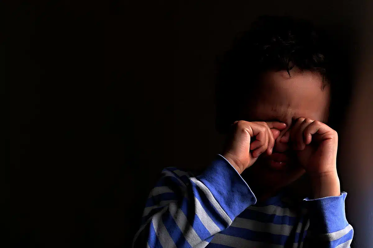 Un petit garçon métisse avec un pull rayé bleu et blanc. Le garçon se tient les yeux car il a peur du noir.
