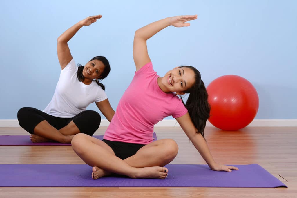 2 jeunes adolescentes qui  pratique de la gymnastique à la salle de gym. Elles sont sur des tapis de sport dans une salle de sport. Il y a un ballon de sport rouge en arrière plan