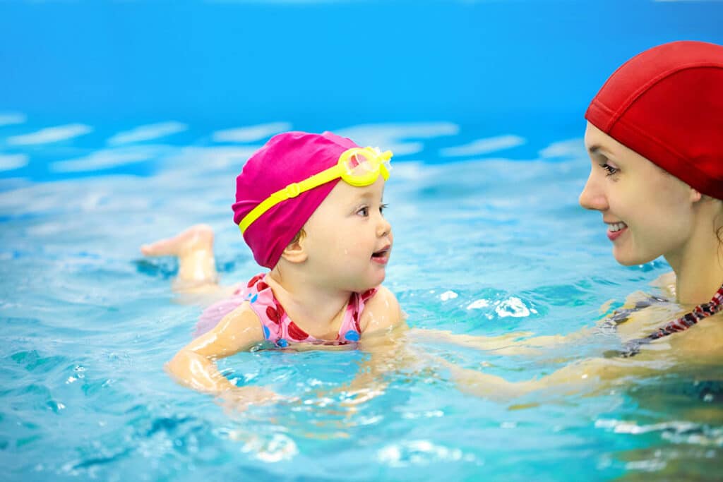 Un bébé fille qui nage dans la piscine pour la première fois avec sa maman. Le bébé porte un bonnet de natation rose et une paire de lunette jaune ainsi qu'un maillot de bain rose. 
