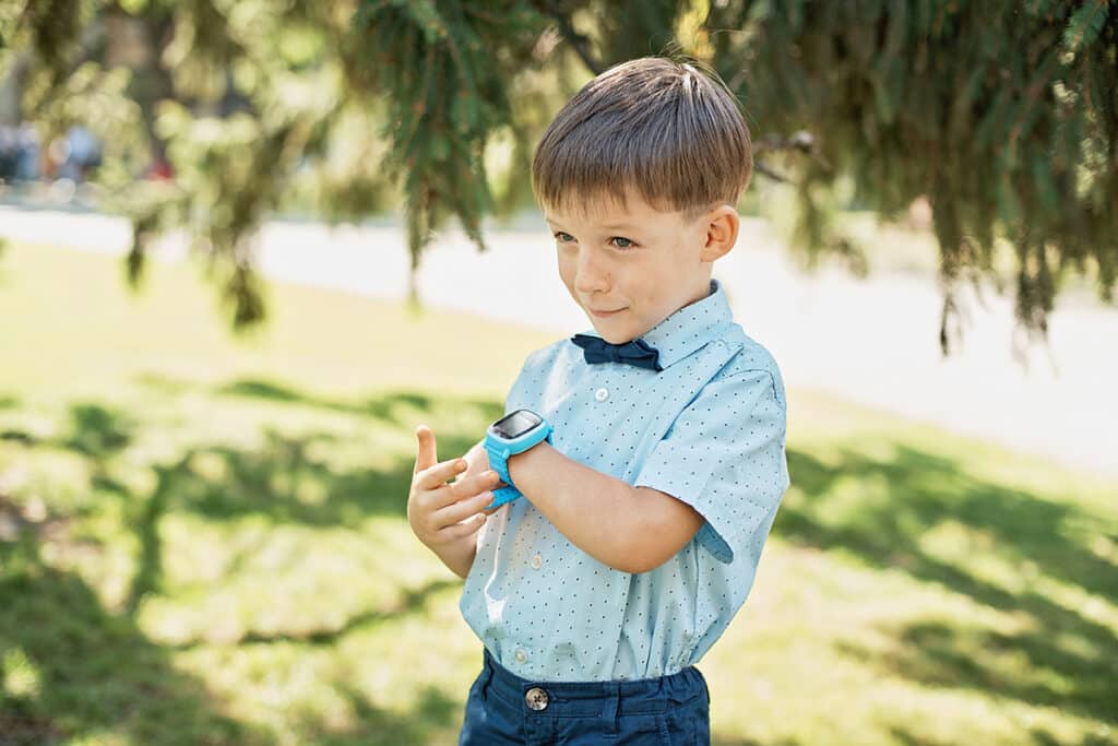Un jeune garçon qui parle dans sa montre connectée. Il porte une montre bleu, une chemise bleu et il est sous le soleil en extérieur. il y a des arbres en arrière plan