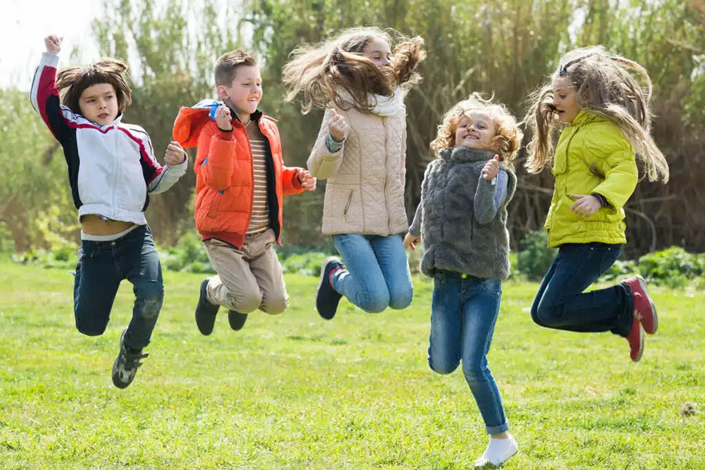 Un groupe de joyeux enfants qui saute dans l'herbe en plein air. Il y a 2 garçons et 3 filles. Il y a des arbres en arrière plan