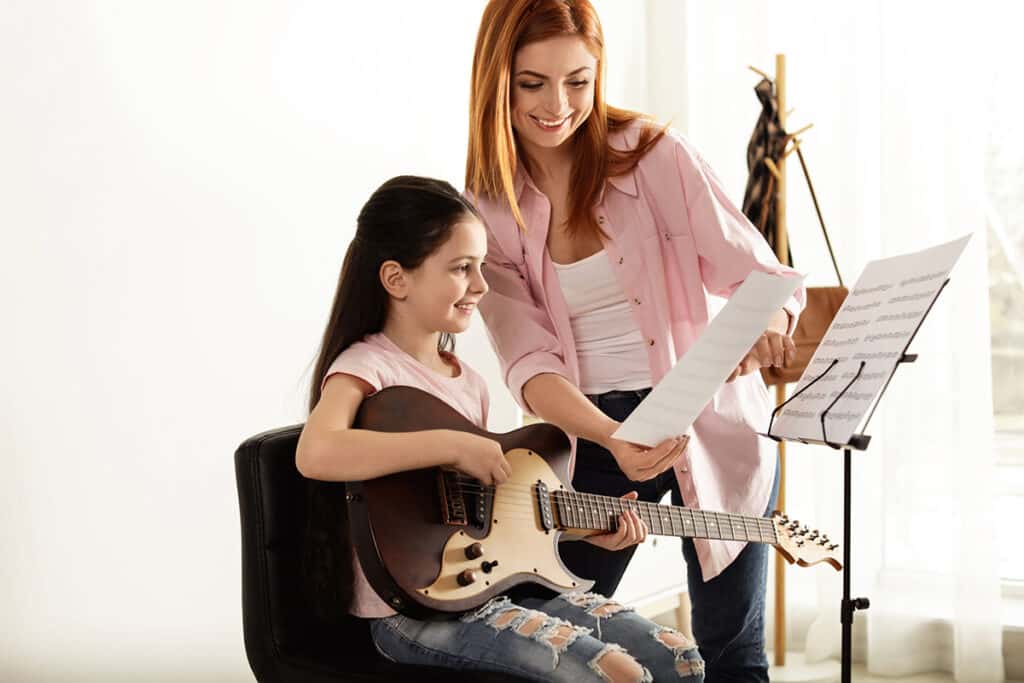 Une jeune fille qui apprend la guitare avec son professeur de musique. La fille porte une guitare éléctrique en bois et est assise face à un chevalet qui tient la partition de musique.