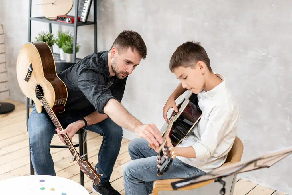 Un moniteur de guitare qui apprend la guitare à un jeune garçon.  Ils sont assis dans un studio de musique, chacun porte une guitare en bois marron