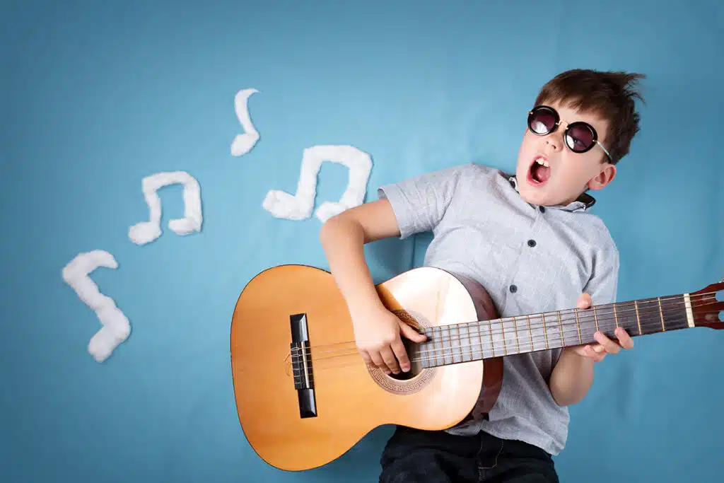 Un jeune garçon sur fond bleu qui fais de la guitare. Le petit garçon porte des lunettes noires, une chemise à courte manche grise et une guitare en bois marron. Il y a des notes de musiques blanches en arrière plan