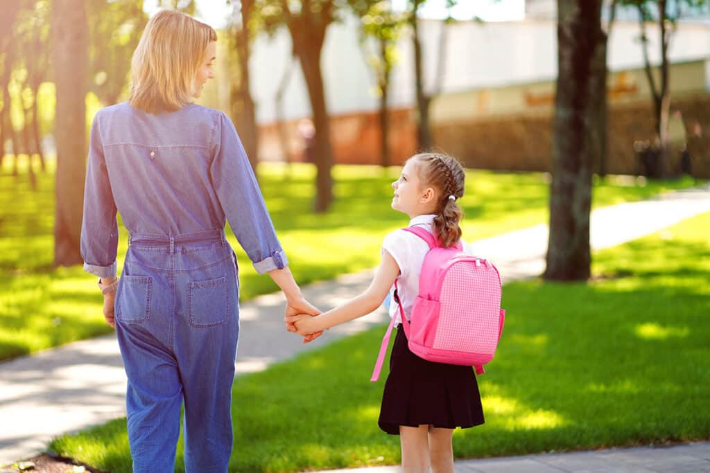 Une mère qui accompagne sa fille dans un parc. Il y a du soleil la mère est habillée en jeans bleu, la petite fille porte un sac à dos pour enfant rose