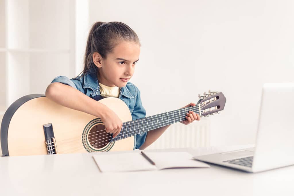 Une jeune fille qui joue de la guitare pour enfant. Elle apprend la guitare devant un cours de musique en ligne sur son ordinateur gris