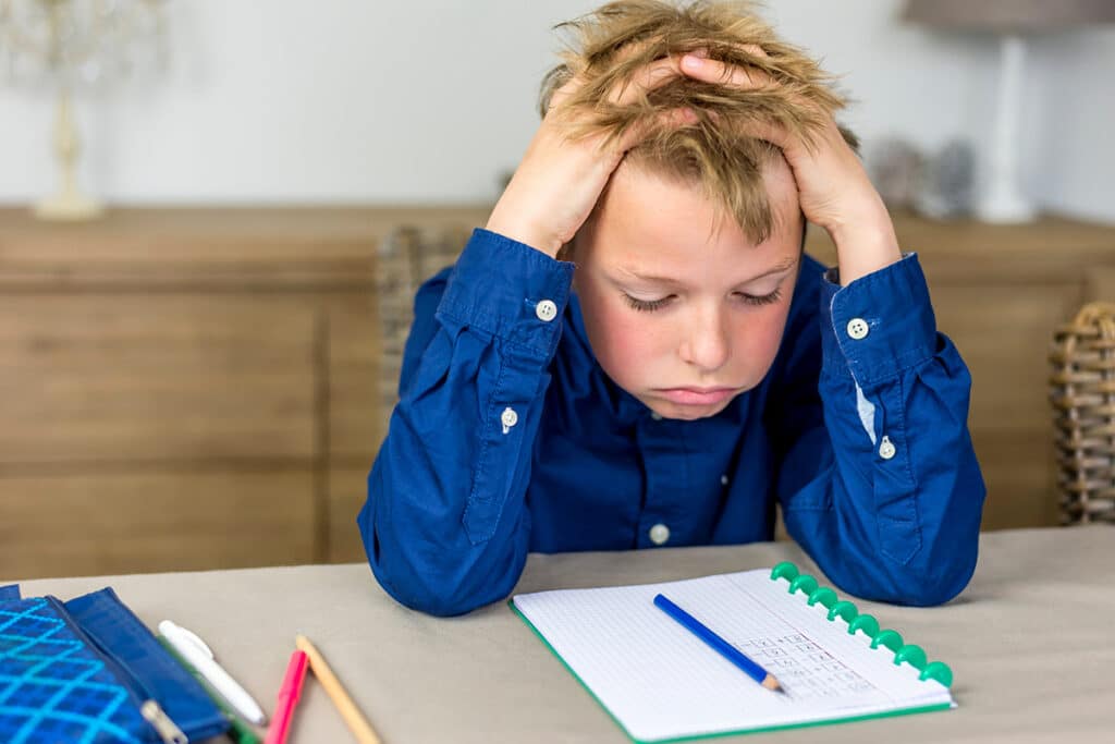 Un jeune garçon en classe d'école assis à table. Il se tient la tête devant son cahier d'exercice. Il est fatigué et porte une chemise pour enfant bleu