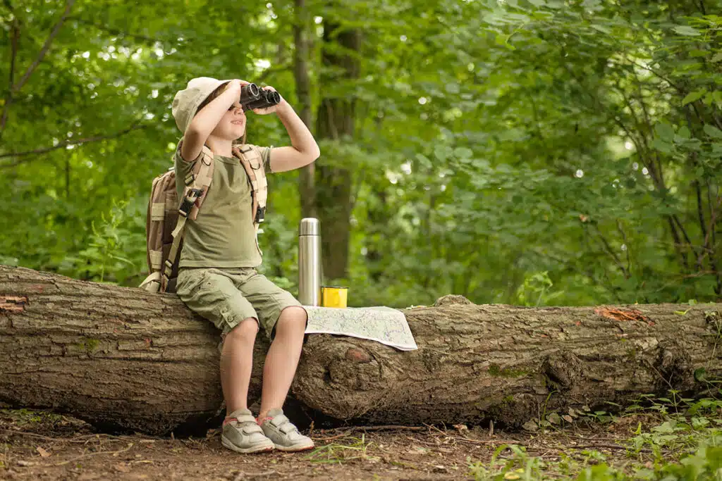 Un jeune garçon en randonnée il est assis sur un arbre et regarde dans ses jumelles. Il a un sac à dos pour enfant et une carte pour le camping