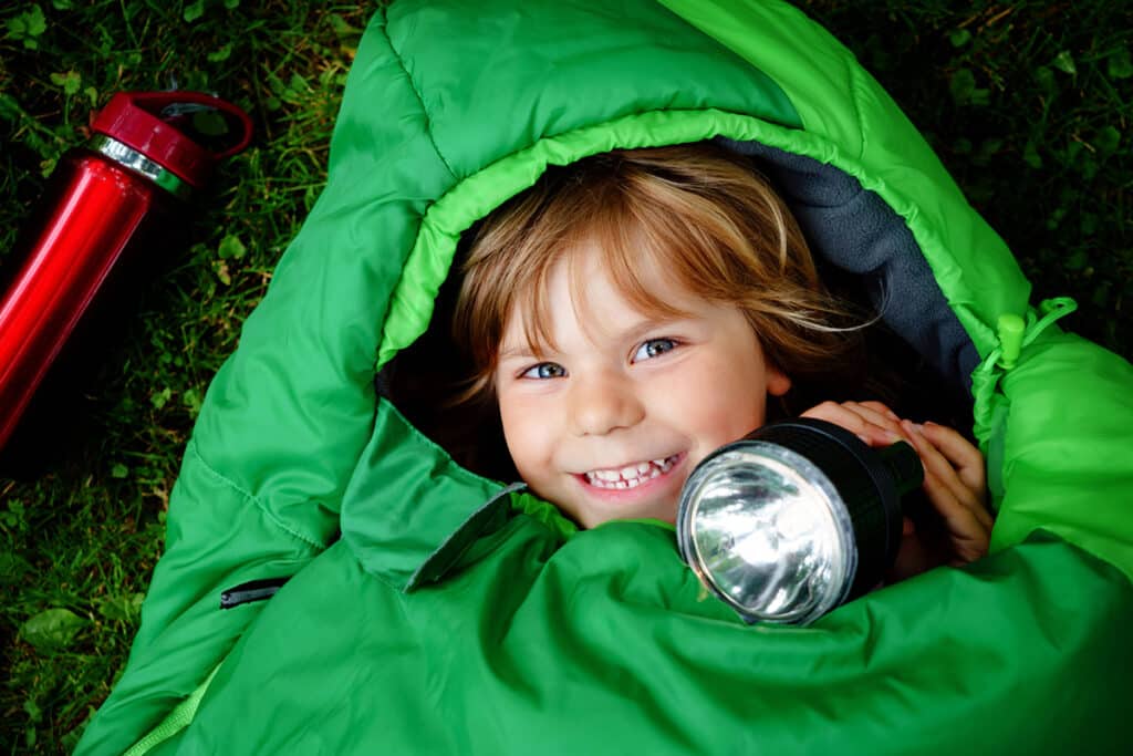 Un jeune garçon roux qui joue dans son sac de couchage pour enfant vert. Il tient une lampe de poche noir 