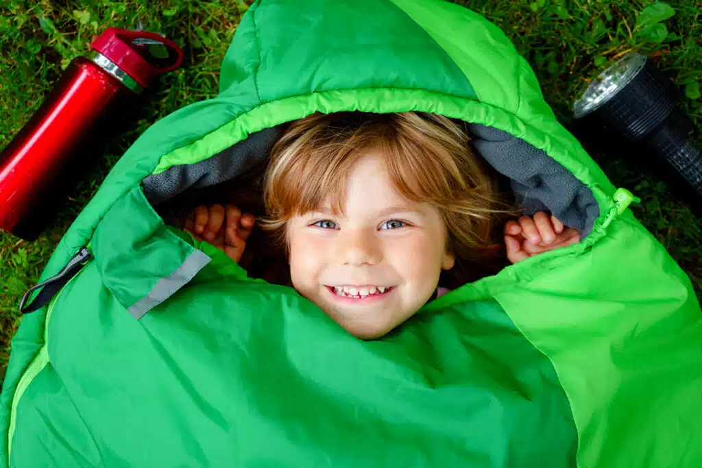 Un jeune garçon roux qui est couché dans son sac de couchage pour enfant. Le sac de couchage est vert. Il y a une gourde à eau rouge et une lampe de poche noire