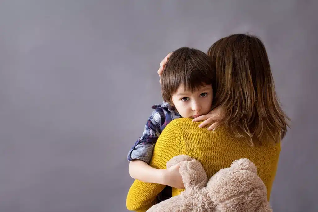 Un petit garçon triste dans les bras de sa maman qui le pardonne. Le petit garçon tient un ours en peluche.