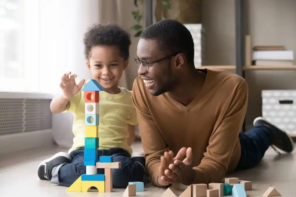 Un mignon petit garçon afro qui joue aux cubes de couleur avec son papa dans leur salon.