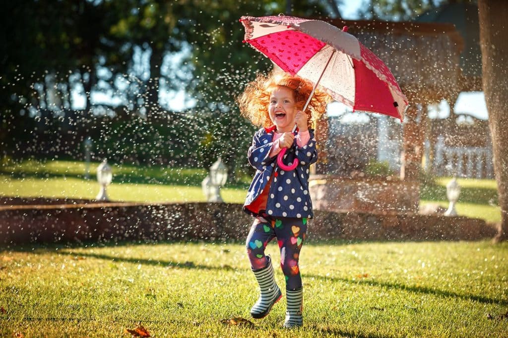 une petite fille  habillée en bleu à pois blanc , au milieu d'un jardin sous le soleil et la pluie. Elle tient un parapluie pour enfant rouge et transparent