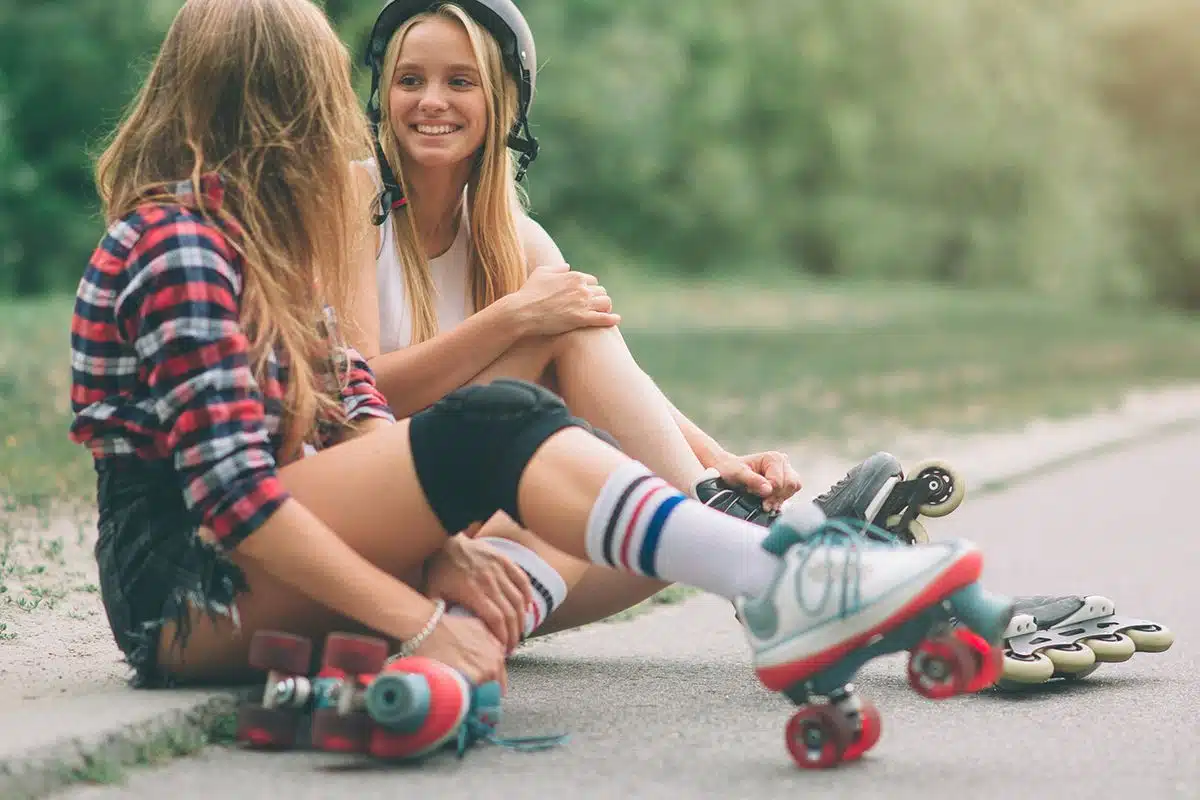 Deux jeunes filles assises sur le bord de la route. Une fille porte des patins à roulettes, l'autre porte des rollers et un casque.