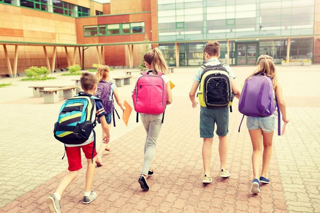 Un groupe de 5 amis qui marchent vers l'école. Il y a deux garçons avec des sacs à dos pour enfant et trois filles avec des cartables d'école.