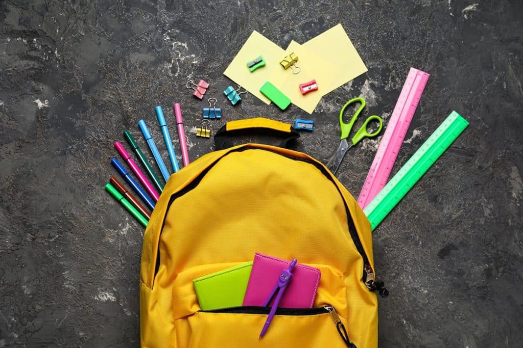 Un sac d'école pour enfant jaune posé sur fond gris. Il y a un compas, des crayons de couleurs, des ciseaux et des règles.