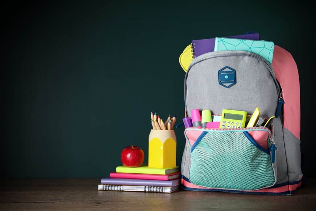 Un cartable d'école en toile grise et rose à demi ouvert. Des cahiers d'école et une calculatrice dépassent du cartable.