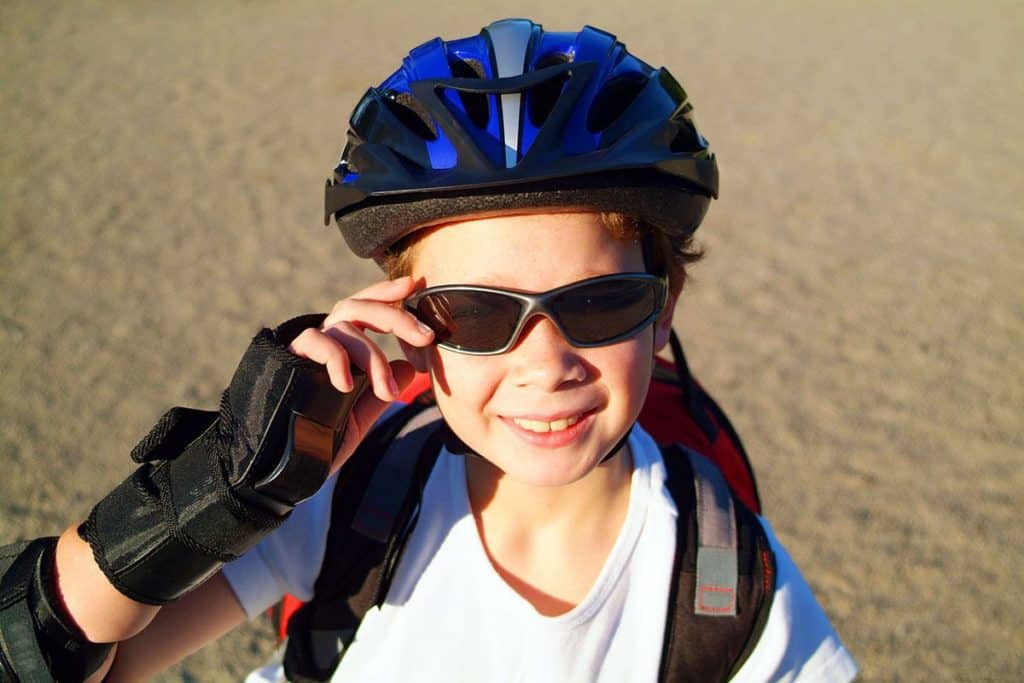 Un jeune garçon qui porte un casque de protection vélo bleu, des lunettes de soleil noires et des gants de protections noir. Il a un sac à dos pour enfant rouge