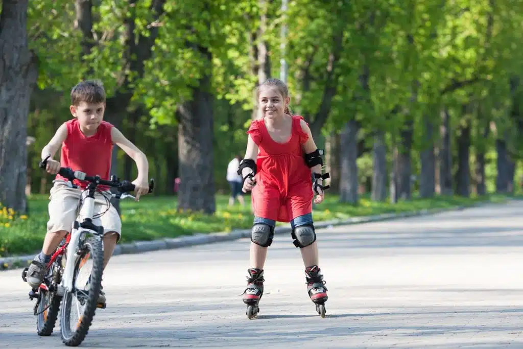 Deux jeunes enfants qui font du vélo et du rollers en toute sécurité. Le garçon est sur un vélo blanc et la fille est sur des rollers rouge et noir. La fille porte des protège-genoux, des protèges-coudes et des gants
