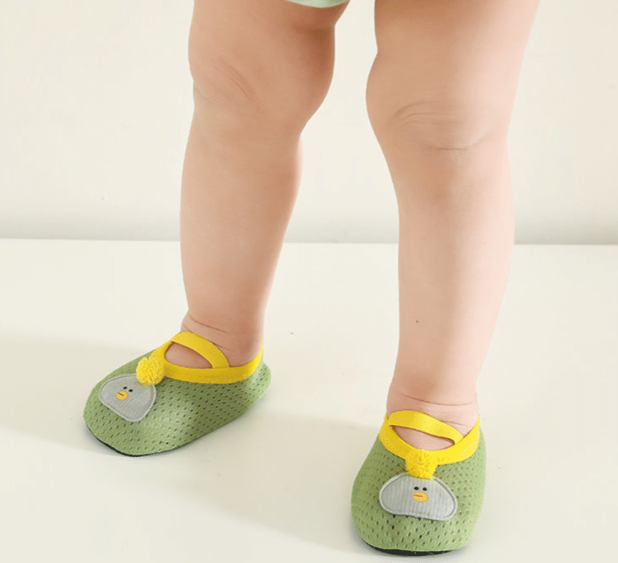 un bébé se tient debout et on ne voit que ses jambes, il porte de petit chaussons verts souples et respirants