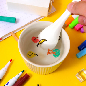 Petit bol blanc rempli d'eau, posé sur un support jaune avec des crayons autour. Une main tient une cuillère et dépose un dessin dans l'eau, grâce à une encre qui flotte