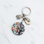 Porte clé en métal avec un rond style vitrail montrant d'un coté mercredi adams en nuance de noir et gris, et de l'autre son amie Enid, colorée. Deux breloques sont accrochées à la chaine : une main manucurée, et la "Chose" , main recousue. Sur fond blanc.
