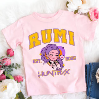 T-shirt enfant K-pop Demon Hunters rose Rumi avec son prénom écrit en majuscules jaune, son visage au centre version cartoon, et Huntrix écrit au-dessous. Le t-shirt est posé à côté de fleurs.