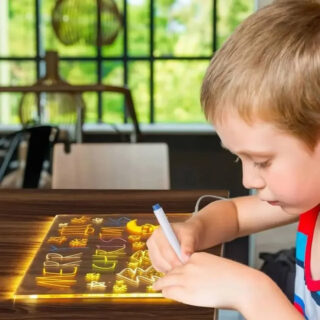 Un enfant dessine avec un stylo fluorescent sur un tableau lumineux LED rétroéclairé avec lettres et formes visibles, dans une ambiance chaleureuse et créative.